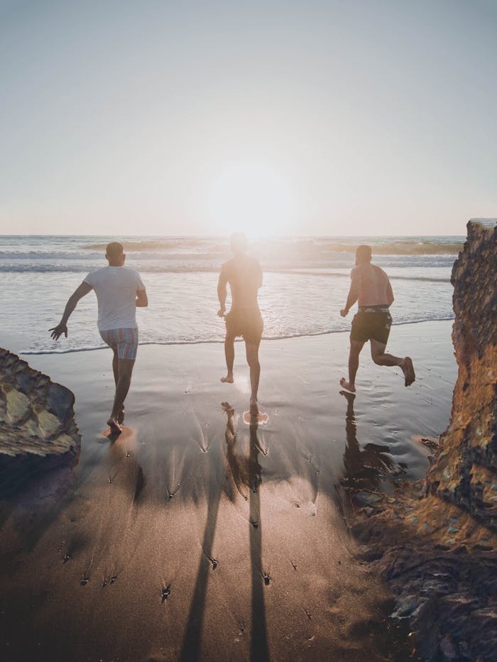 Three men run joyfully towards the ocean at sunset on a sandy beach, enjoying the outdoors.