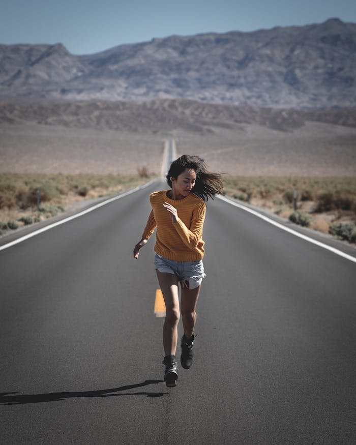 An Asian woman runs confidently on a long, remote desert highway under clear blue skies.